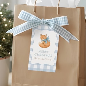May include: A brown paper gift bag with a light blue gingham ribbon and tag. The tag features a watercolor fox illustration and the words "Merry Christmas". A small potted evergreen tree and books are in the background.