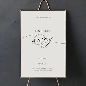 May include: A framed sign on a gold easel with the text "Our Wedding Is One Day Away." The sign also includes the names "Elouise and Beckett" and details for "The Rehearsal Dinner."
