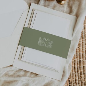 May include: A white invitation suite featuring a sage green belly band with a white line drawing of two swans facing each other. The set includes a matching white envelope and a round wooden wax seal stamp with a botanical leaf design.