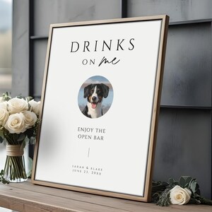 May include: A framed sign with the text "DRINKS on me" and a photo of a dog. Below the photo, it reads "ENJOY THE OPEN BAR". The sign is on a wooden table with a bouquet of white roses.