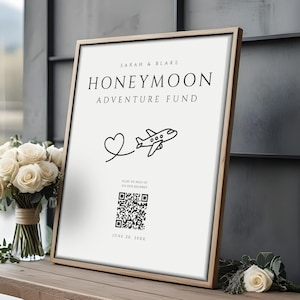 May include: A framed sign with the text "Sarah & Blake HONEYMOON ADVENTURE FUND" and a heart and airplane graphic. The sign is on a wooden table with a bouquet of white roses and a single rose. The date "JUNE 26, 20XX" is at the bottom.