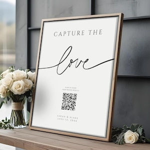 Op de afbeelding: Een ingelijst bord met de woorden "CAPTURE THE love" in zwart schrift. Daaronder een QR-code, de namen "SARAH & BLAKE" en een datum. Een boeket witte rozen staat links op een houten bank.