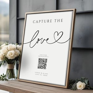 May include: A framed sign with the words "CAPTURE THE love" in black script. Below the text is a QR code and the names "SARAH & BLAKE" and the date "JUNE 12, 20XX". The sign is on a wooden table with a bouquet of white roses.
