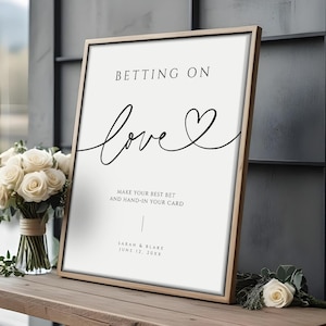 May include: A framed sign with the words "Betting on Love" in black script. The sign includes the text "Make your best bet and hand-in your card" and the names Sarah & Blake, June 12, 20XX. A bouquet of white roses sits to the left.