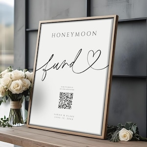 May include: A framed sign with the words "Honeymoon Fund" in black script. Below the text is a QR code and the names "Sarah & Blake" with the date "June 12, 20XX". The sign is on a wooden bench with a bouquet of white roses.