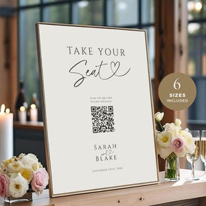 May include: A framed sign with the text "TAKE YOUR Seat" in elegant script. The sign includes a QR code and the names "SARAH and BLAKE" with a wedding date. A small bouquet of flowers and champagne glasses are on the table.