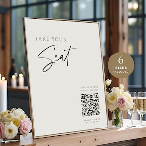 May include: A framed sign with the text "TAKE YOUR Seat" in elegant script. The sign includes a QR code and the names "SARAH - BLAKE" with a date. A small bouquet of flowers and champagne glasses are on the wooden table.