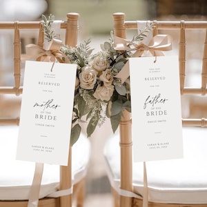 Könnte beinhalten: Zwei Holzstühle mit weißen Kissen, die für eine Hochzeit dekoriert sind. Jeder Stuhl hat ein Schild mit der Aufschrift „Reserviert“ und weist Sitzplätze für die „Mutter der Braut“ und den „Vater der Braut“ aus. Blumengestecke und Bänder schmücken die Stühle.