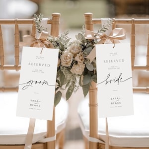 Könnte beinhalten: Dekoration für Hochzeitszeremonie-Stühle. Zwei Holzstühle mit weißen Kissen sind mit Blumengestecken und Schildern mit der Aufschrift "Reserviert" und den Namen "Groom" und "Bride" geschmückt. Die Schilder sind mit Bändern befestigt.