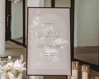 May include: A black easel with a white sign that says "Welcome to Olivia's Bat Mitzvah, Saturday 14 December, Long Island New York". The sign is on a white table with candles in glass cylinders.
