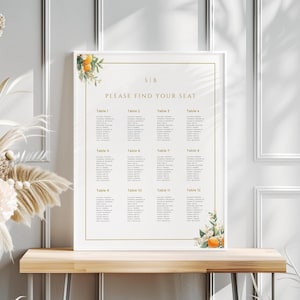 May include: A white framed seating chart with a floral design featuring orange blossoms and green leaves. The chart is titled "Please Find Your Seat" and lists tables with names for each guest.