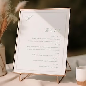 Może przedstawiać: Złota ramka z listą koktajli, win, piw, likierów i napojów gazowanych. Znak znajduje się na białym obrusie, z wazonem z suszonymi kwiatami i małym kubkiem w tle.