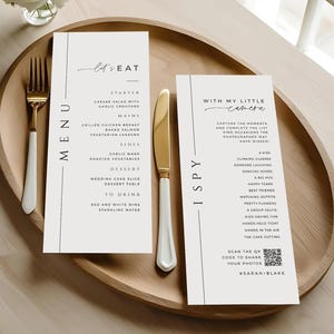 May include: A wedding menu and an "I Spy" game card are displayed on a wooden tray, alongside gold cutlery. The menu lists courses like Caesar salad and grilled chicken. The "I Spy" card includes prompts like "clinking glasses" and "dancing shoes".