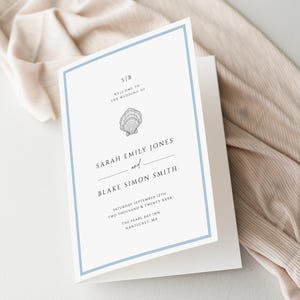 Könnte beinhalten: Eine gefaltete Hochzeitseinladung mit hellblauem Rand. Die Einladung zeigt die Namen "Sarah Emily Jones" und "Blake Simon Smith" mit dem Text "Willkommen zur Hochzeit von" und einer Muschelgrafik. Datum und Ort sind ebenfalls aufgedruckt.
