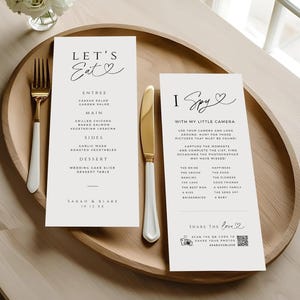May include: A wooden tray with wedding stationery, including a menu and an "I Spy" game card. The menu lists entree, main, sides, and dessert options. A gold fork and knife with white handles are on the tray, with a small vase of white flowers.