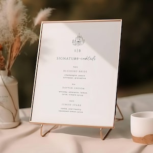 May include: A gold-framed sign with a white background and black text lists signature cocktails for a wedding. The sign reads "SB SIGNATURE cocktails" and lists the drinks "Blushing Bride" and "Dapper Groom" with their ingredients.