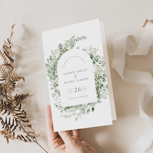 May include: A white wedding invitation with a green watercolor wreath featuring leaves and flowers. The text reads "Welcome to the wedding of Sarah Emily & Blake Simon Sat 26 Aug Toronto, Ontario".