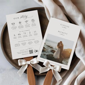 May include: Two wedding fan programs with cream paper and wooden handles, decorated with white ribbon bows. One has text detailing the couple's story, the other a photo of the couple. The text includes the names Elouise and Beckett.