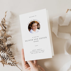 Puede incluir: Una tarjeta blanca con texto negro anunciando una Bar Mitzvah para Oliver Leo Silver. La tarjeta incluye una foto del niño en un círculo. El texto dice "Bienvenido a la Bar Mitzvah de nuestro hijo Oliver Leo Silver" en hebreo e inglés. La fecha, la ubicación y el templo se enumeran a continuación.