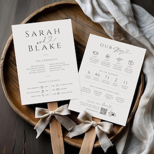 May include: Two wedding ceremony signs with the names "Sarah and Blake" and "Our Story" printed in black on off-white paper. The signs are on wooden sticks with cream-colored bows. The signs are displayed on a wooden tray.
