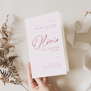 May include: A white card with pink text announcing a Bat Mitzvah for Olivia Eve Silver. The card includes the date, location, and the Hebrew transliteration of Olivia's name.