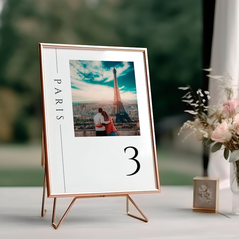 K&ouml;nnte beinhalten: Eine goldene Tischkarte mit einem Foto vom Eiffelturm und einem Paar in Paris. Die Nummer "3" ist unter dem Foto gedruckt. Der Rahmen steht auf einem goldenen Metallst&auml;nder.