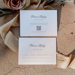 May include: Two white RSVP cards with elegant script that reads "Please Reply." The cards include prompts for accepting or declining, space for names, and a QR code for more details. The cards are on a textured surface.