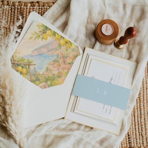 May include: Wedding stationery set with a watercolour landscape design inside the envelope. The set includes invitation cards, a pale blue belly band with initials, and a wooden wax seal stamp. The colour scheme is soft and inviting.
