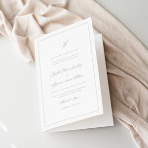 May include: A folded white wedding program with a grey border, featuring elegant script and the names of the couple and their parents. The background is a beige fabric. The program is designed for a wedding ceremony.