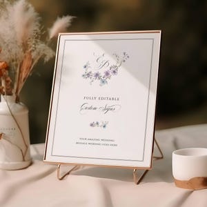 May include: A framed sign with a gold metal stand. The white sign has a floral design with purple and blue flowers and the text "FULLY EDITABLE Custom Signs". The sign is on a white surface.