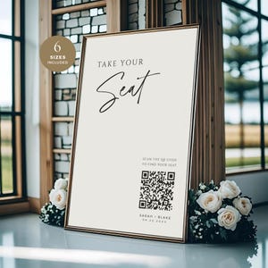 Puede incluir: Un letrero enmarcado con el texto "TAKE YOUR Seat" en escritura elegante. El letrero incluye un código QR y los nombres "SARAH + BLAKE" con una fecha. El letrero está rodeado de rosas blancas y vegetación, adecuado para una boda o evento.