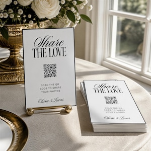 May include: A white rectangular sign with a black border on a gold stand, featuring the text Share the Love, a QR code, instructions to scan to share photos, and the names Olivia & Lucas. A stack of identical cards sits next to the sign.