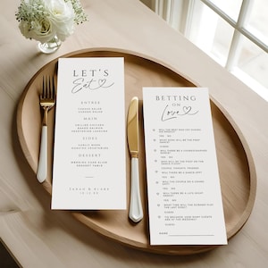 Puede incluir: Un juego de menú y tarjetas de boda en una bandeja de madera. El menú dice "Let's Eat" con una lista de entrantes, platos principales, guarniciones y postres. La tarjeta del juego se titula "Betting on Love" con preguntas.