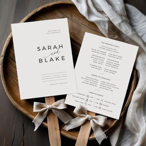 May include: Two wedding program fans with beige bows rest on a wooden tray. The fans display the names "Sarah and Blake" and wedding details in black text. The tray is made of dark wood, and a white cloth is in the background.