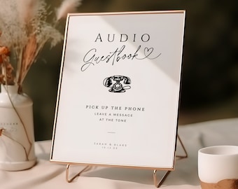 May include: A framed sign with the text "AUDIO Guestbook" and a vintage telephone illustration. The sign is in a rose gold frame and stand, with instructions to leave a message. The sign is on a table with a white vase and a small cup.