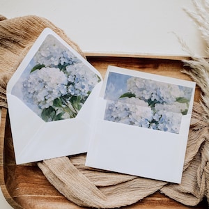 Può includere: Due buste bianche con un motivo floreale. Le buste sono aperte, rivelando un dipinto ad acquerello di ortensie blu e bianche. Le buste sono su un vassoio di legno con un tessuto beige.