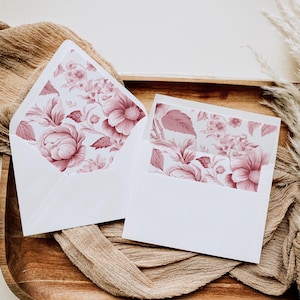 May include: Two white envelopes with a floral pattern in shades of pink. The envelopes are on a brown surface.