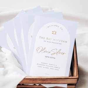 Puede incluir: Una pila de tarjetas azul claro con una estrella dorada y el texto "Bienvenido a la Bat Mitzvah de nuestra hija Olivia Silver" en un arco blanco. El texto "אוליבה סילבר" también está incluido en hebreo. Las tarjetas también incluyen la fecha, la ubicación y la hora del evento.