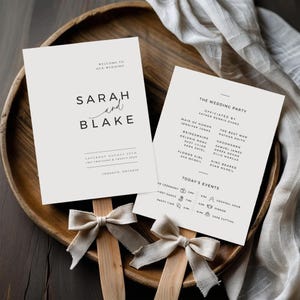 Może przedstawiać: Dwa programy weselne z tekstem „SARAH and BLAKE” i „THE WEDDING PARTY” spoczywają na drewnianej tacy. Programy są jasne z czarnym tekstem. Do programów przymocowane są drewniane patyczki z kremowymi kokardkami.