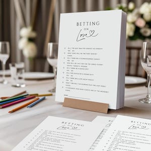 Puede incluir: Una pila de tarjetas blancas para juegos de boda con el texto "Betting on Love" en escritura negra. Las tarjetas están sujetas por un tarjetero marrón. Lápices de colores y copas de vino en el fondo.