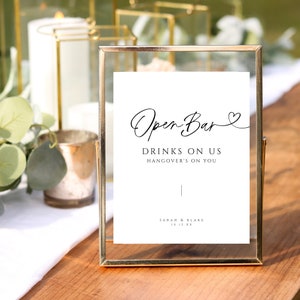 May include: A gold-framed sign with the text "Open Bar" in cursive script, followed by "Drinks on us, hangover's on you". The sign also includes the names "Sarah & Blake" and the date "10.12.XX".