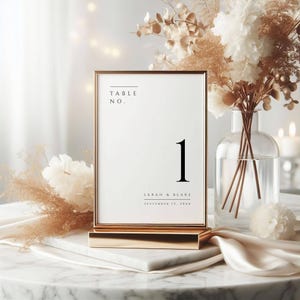 May include: Gold table number card with a white background and black text. The card reads "Table No. 1" and "Sarah & Blake September 12, 20XX".