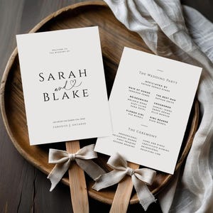 Puede incluir: Dos abanicos de programa de boda con texto "SARAH and BLAKE" y "THE WEDDING PARTY". Los abanicos son de color beige claro con texto negro y están decorados con lazos beige. Los abanicos están en una bandeja de madera.