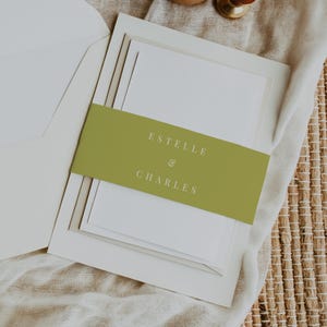 May include: A wedding invitation suite featuring a stack of white cards with a green belly band. The band displays the names "Estelle & Charles" in a simple, elegant font. The suite is presented with a white envelope.