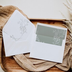 May include: A white envelope with a delicate floral design in grey. The envelope is open to reveal a smaller, grey envelope with a similar floral design. The envelopes are on a brown wooden surface with a beige fabric.