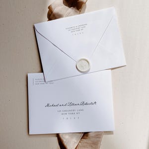 May include: Two white envelopes with elegant calligraphy and addresses. One envelope is sealed with a floral wax stamp. The text on the envelopes includes names, street addresses, and postcodes. The envelopes are likely for wedding invitations or announcements.