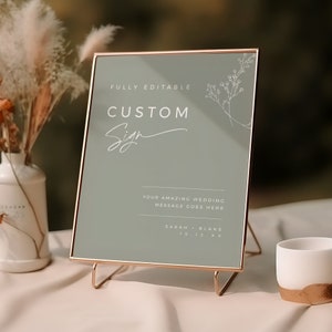 Może przedstawiać: Szklana tablica w kolorze szałwii z pozłacanym ramą i stojakiem. Na tablicy widnieje napis "FULLY EDITABLE CUSTOM Sign" w ozdobnym, ręcznie pisanym stylu. Pod tytułem znajduje się tekst "YOUR AMAZING WEDDING MESSAGE GOES HERE". Na dole tablicy widnieją imiona "SARAH + BLAKE" oraz data "10.12.XX".