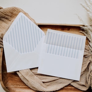 May include: Two white envelopes with blue striped lining are displayed open on a wooden tray. The envelopes are positioned on a beige fabric, with a neutral background. The envelopes are likely for invitations or stationery.
