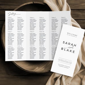May include: A wedding seating chart and welcome sign. The seating chart lists guests by table number, with names in black text on a white background. The welcome sign says "Welcome to the Wedding of Sarah and Blake."