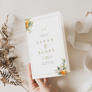 Puede incluir: Una invitación de boda blanca con letras doradas y un diseño floral con flores de azahar y cítricos. La invitación dice "Bienvenido a la boda de Sarah & Blake, Sáb 26 Ago, el resort tropical, Palm Beach, Florida."